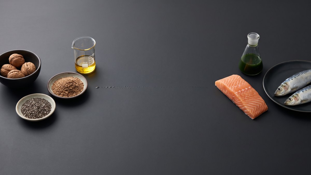 On the left walnuts, ground flaxseed, chia seeds and a beaker of light oil; on the right a salmon fillet, two sardines and a flask of green microalgae culture; between them a fine, fading trail of droplets on a dark table