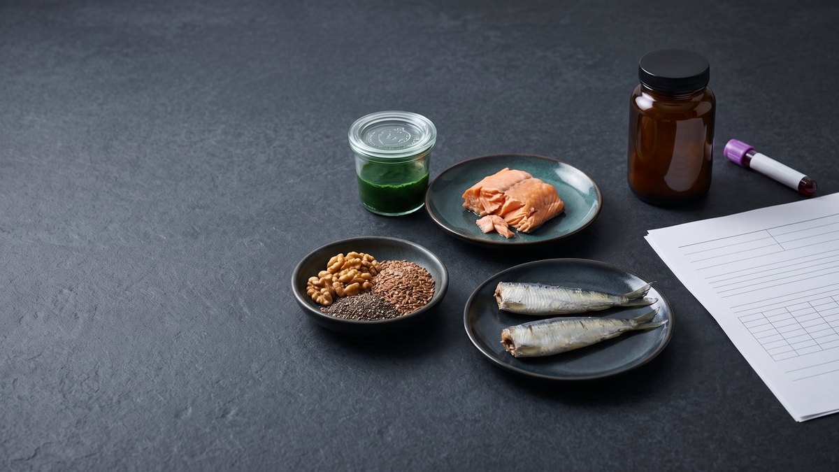 A piece of salmon, two sardines, a bowl of walnuts, flaxseed and chia seeds and a jar of green microalgae culture next to a closed amber glass jar, a blood sample tube and laboratory sheets on a dark surface